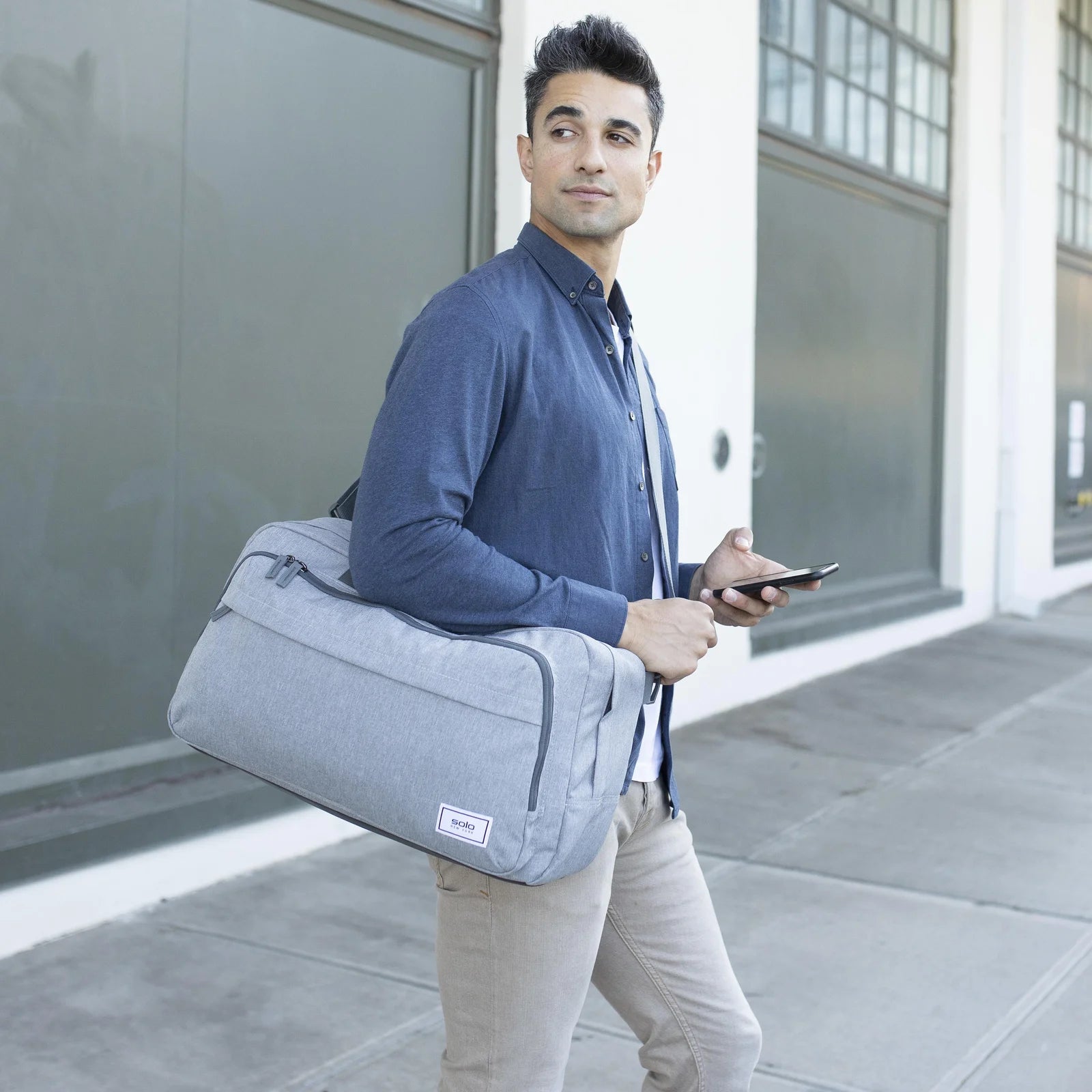 גבר ברחוב עירוני נושא תיק דאפל SOLO New York דגם Re:Move מבד ממוחזר בצבע אפור ביד - תיק סוף שבוע בעיצוב עסקי נקי